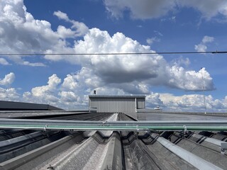 Close-up low-angle view of a corrugated metal rooftop, focusing on an electrical conduit system and a large metal structure against a dramatic blue sky with white cumulus clouds.