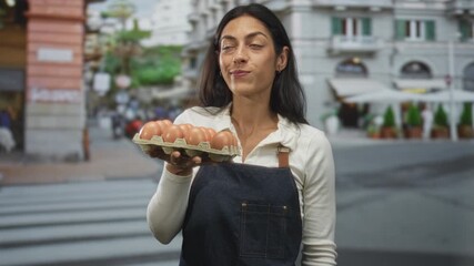 Woman holds egg carton of brown eggs with one hand at chest level, wearing denim apron and smiling in front of a street cafe; local market pride.