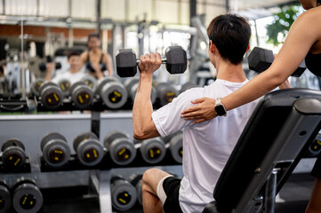 Man client holding dumbbell exercising and fitness trainer helping fixing posture across gym mirror.