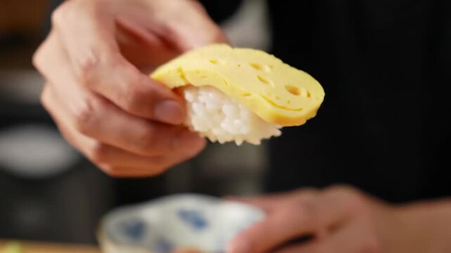 Hand holds tamago nigiri, a rectangle of egg atop rice. Soft focus shows a blurred background