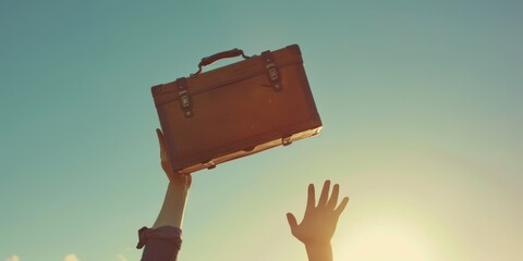 A person's hand holding a suitcase against a clear blue sky with the sun shining through.