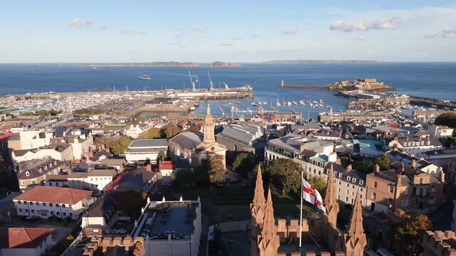 Flight over Elizabeth College St James and rooftops of St Peter Port Guernsey towards harbour Herm Sark and Jethou in the distance in bright afternoon sunshine on calm day