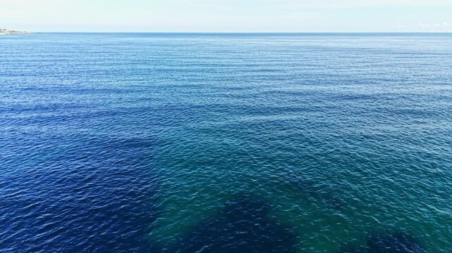 Calm waters shimmer under the sun, creating a peaceful scene. Gentle waves roll softly, while a few distant landforms break the horizon, enhancing the sense of serenity.