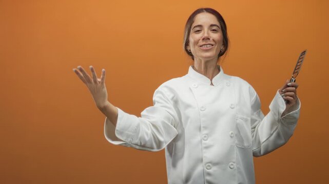 Young woman chef in white coat holds metal grater in right hand while raising left hand palm up in a tasting gesture against orange backdrop in studio; confidence.