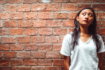 Southeast Asian teenage girl with long black hair in white shirt standing in front of a brick wall background looking up to the light and thinking