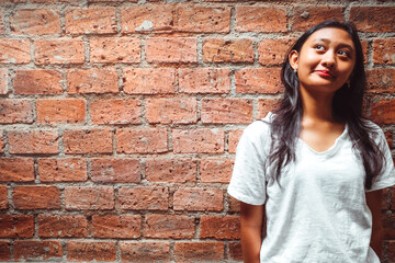 Asian teenage girl with long black hair in white shirt standing in front of brick wall looking up to the light and smiling