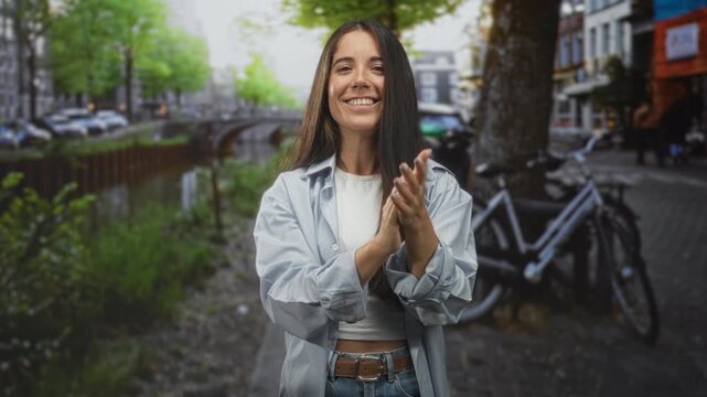 Young hispanic woman clapping hands and smiling, bare midriff visible, standing on a street beside a canal with parked bicycle and trees; joy celebration.