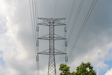 A large power line tower or electrical pylon against a dramatic cloudy sky and green trees below. Concepts of energy, electricity, infrastructure, global warming, environment, and industrial silhouett