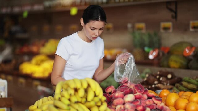 Woman near vegetable stand in store choose flat peach. Client view ripeness of fruit, take out fruits from showcase, collects grocery basket. Girl pull out ripest fruits from large pile of fruits