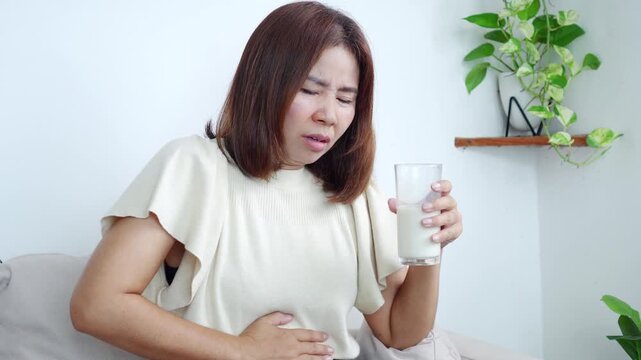 Asian woman stomach ache after drinking milk caused by lactose intolerance or digestive discomfort. Concept of dairy sensitivity, upset stomach, bloating, and food-related health problems. 