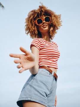Bold sunlit portrait of a stylish woman reaching toward the camera, wearing a red and white striped crop top, high waisted denim shorts, and oversized round sunglasses against a clear blue sky.