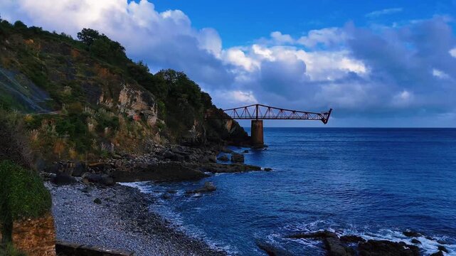 Approche lente d'un pilonne de b&eacute;ton surplomb&eacute; par une structure m&eacute;tallique au bord d'une c&ocirc;te rocheuse. Vue de l'oc&eacute;an au drone, ciel bleu. Format horizontal 16:9 - 4K 