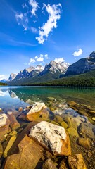 A vibrant scenic view features calm water, rocky foreground, mirroring tall snow-capped mountains and lush green trees beneath a bright blue sky