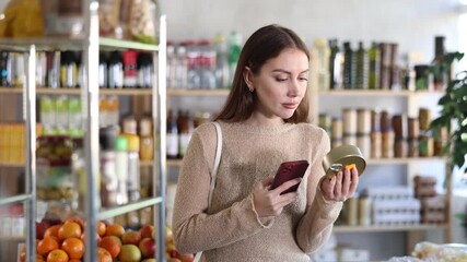 Thoughtful young woman making purchases in local grocery store, using smartphone to scan barcode on canned food packed in tin can. Modern shopping concept