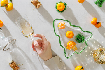 Top view festive composition with tangerines and glass rose wine in woman hand, green glass Christmas tree shaped  tray, champagne corks, discoball. Long shadow, Christmas and New Year holidays mood.