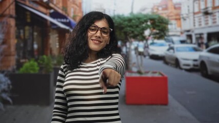 Woman pointing finger on street wearing glasses and striped shirt, direct gaze and lip piercing while reaching forward; bold confidence.