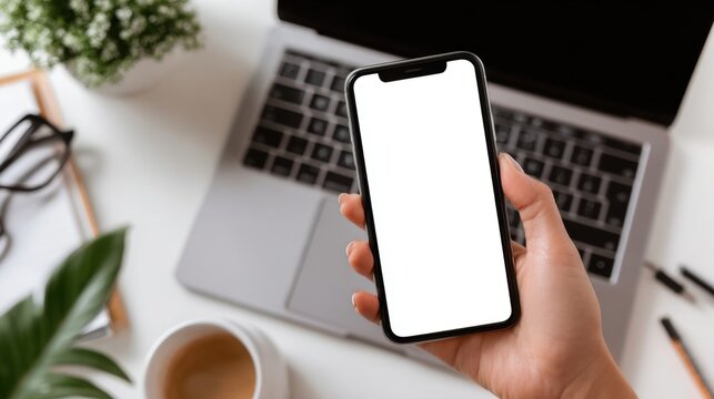 Hand holding a blank screen smartphone near a laptop coffee cup and glasses