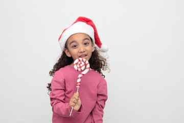 4-year-old Latina girl with curly brown hair wearing a santa hat sings with a candy cane to celebrate Christmas and New Year