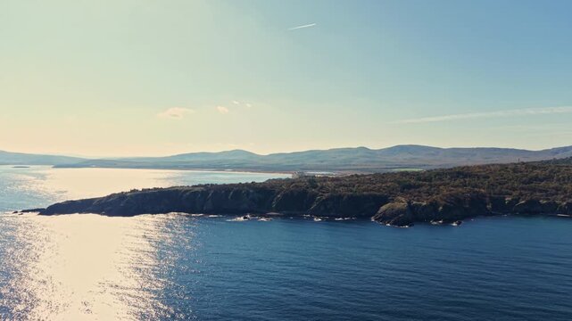 Gentle morning light dances on the peaceful bay, illuminating rocky shores and distant hills under a clear blue sky with wisps of clouds.