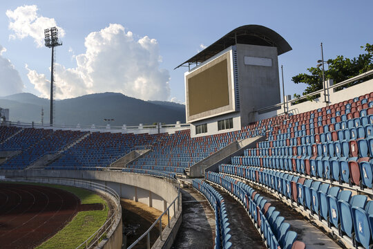 Quiet outdoor sports arena with rows of empty blue and red seat and large scoreboard. grandstand feels peaceful under partly cloudy sky with mountain view - Powered by Adobe
