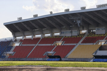 Obraz premium Quiet, empty stadium showing colorful red, blue, and yellow seats under large roof. view from green athletic field captures grandstand with feeling of anticipation