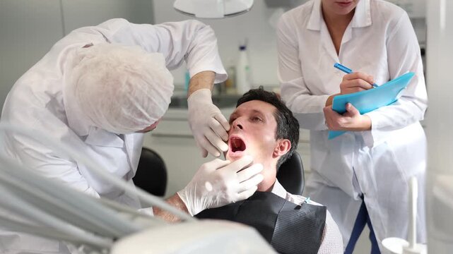 Elderly male doctor dentist with young female nurse examining teeth of male patient before treatment in office