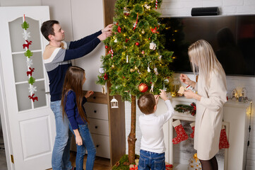 Happy family decorates home together during Christmas holidays. Mom, Dad, brother, sister happily decorate Christmas tree in cozy home and have fun, creating warm, festive atmosphere.