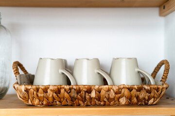 Aesthetic Home Organization. Three Ceramic Mugs Stored Upside Down in a Natural Woven Hyacinth Basket on a Wooden Shelf