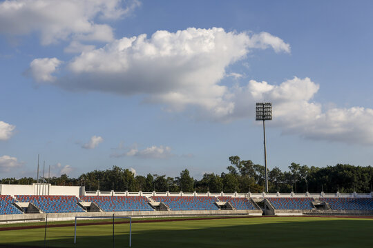 Calm empty sport stadium featuring green field and blue seats under blue sky with white cloud. Quiet outdoor arena awaits future game. Peaceful athletic venue scenery