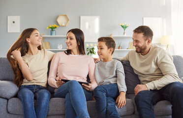 Happy family of four sitting on sofa in living room, enjoying warm and cheerful conversation and laughter. Parents and children spending quality time together, having fun and sharing comical stories.