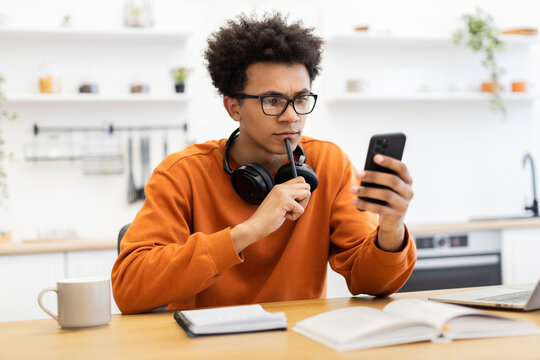 A young man with glasses and headphones looks at his phone thoughtfully while sitting at a table with a notebook and coffee cup.