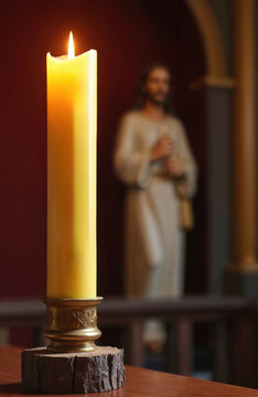 Tall yellow Paschal candle burns brightly in church with Jesus statue in background. Lighted wax candle on wooden base, symbol of Easter celebration and faith.