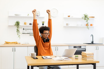 A young man celebrates with arms raised, looking at a laptop in a bright kitchen setting.