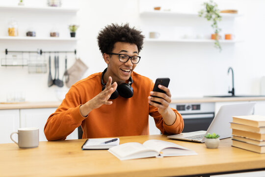 A young man with glasses is smiling and gesturing while on a video call using his smartphone at his kitchen table.