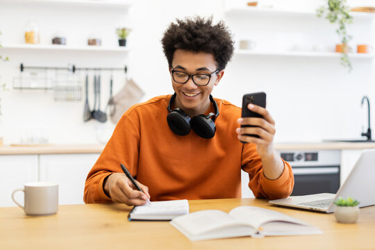 A young man with glasses smiles while looking at his phone and writing in a notebook at a table with a laptop and coffee cup. - Powered by Adobe