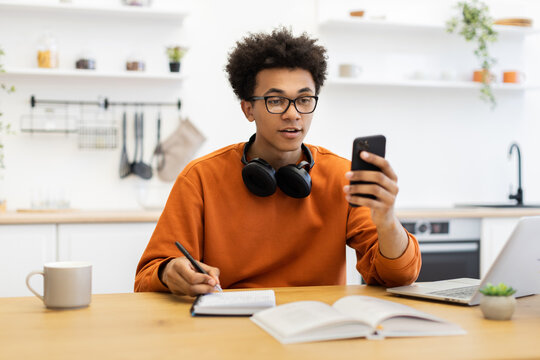 A young man with glasses is using his phone while working at a table with a laptop and notebook.