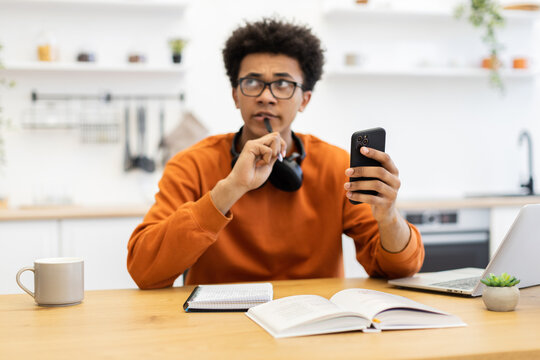 A young man in glasses contemplates while holding a phone and pen, with a book and laptop on the table. - Powered by Adobe