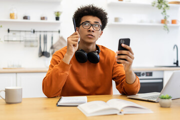 A young man contemplates while holding a pen and smartphone, possibly working from home in a kitchen setting.