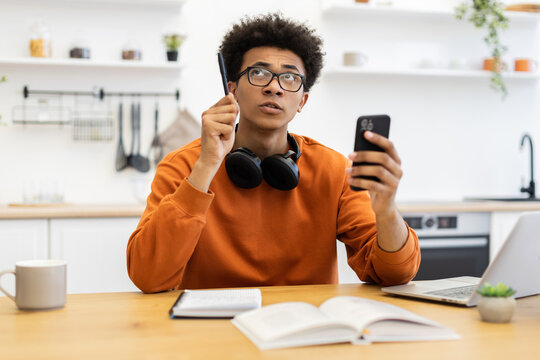 A young man contemplates while holding a pen and smartphone, possibly working from home in a kitchen setting. - Powered by Adobe