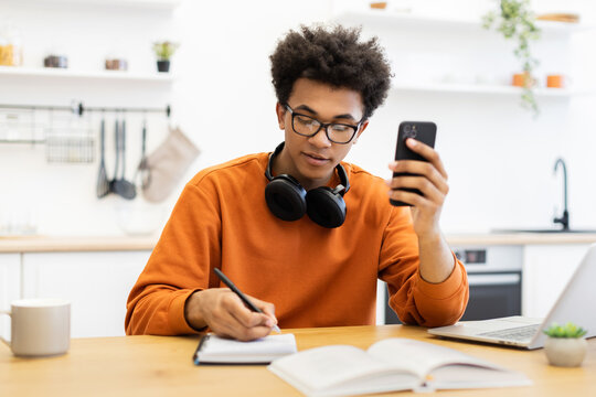 A young man with glasses is working at a table, using his phone and writing in a notebook. - Powered by Adobe