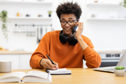 A young man with glasses is on the phone while taking notes at his desk, with a laptop and coffee cup nearby. - Powered by Adobe