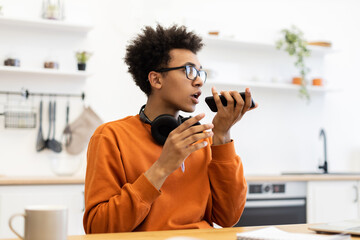 A young man with glasses speaks into his phone, possibly using a voice assistant, in a bright kitchen setting.