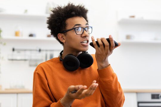 A young man with glasses speaks into his smartphone, likely using a voice assistant or making a call, with headphones around his neck.