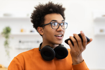 A young man with glasses and headphones is speaking into his phone, possibly using a voice assistant.