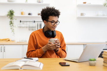 A young man with glasses enjoys a cup of coffee while working on his laptop at home.