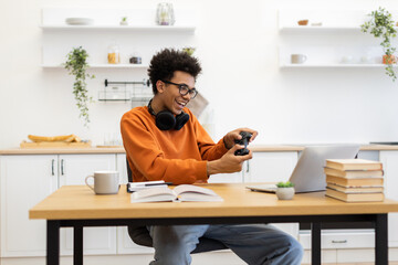 A young man with glasses is smiling while playing video games on his laptop at a wooden table.