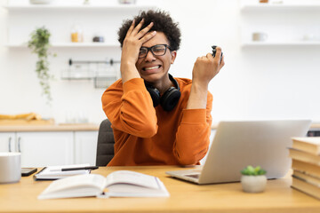 A young man wearing glasses looks stressed while holding a game controller and sitting at a desk with a laptop and books.