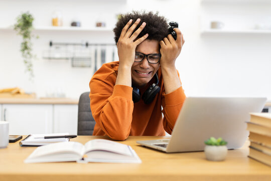 A young man is visibly upset, holding a game controller, and looking at a laptop screen.