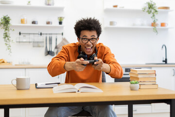 A young man with glasses is enthusiastically playing a video game in his kitchen, holding a controller with headphones around his neck.