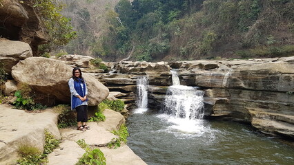 Woman standing by a Nafakhum Waterfall in a natural setting on a sunny day with flowing water and...